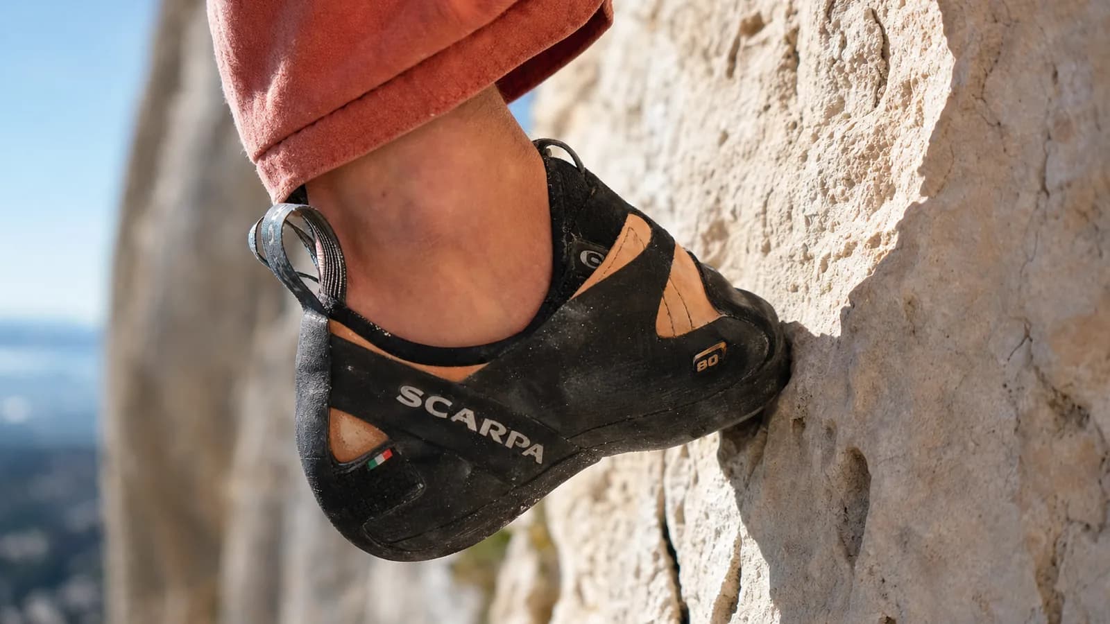 Plan rapproché d'un chausson rouge La Sportiva posé en pointe sur une micro prise de granite gris, gomme noire écrasée contre le rocher, magnésie blanche sur le chausson