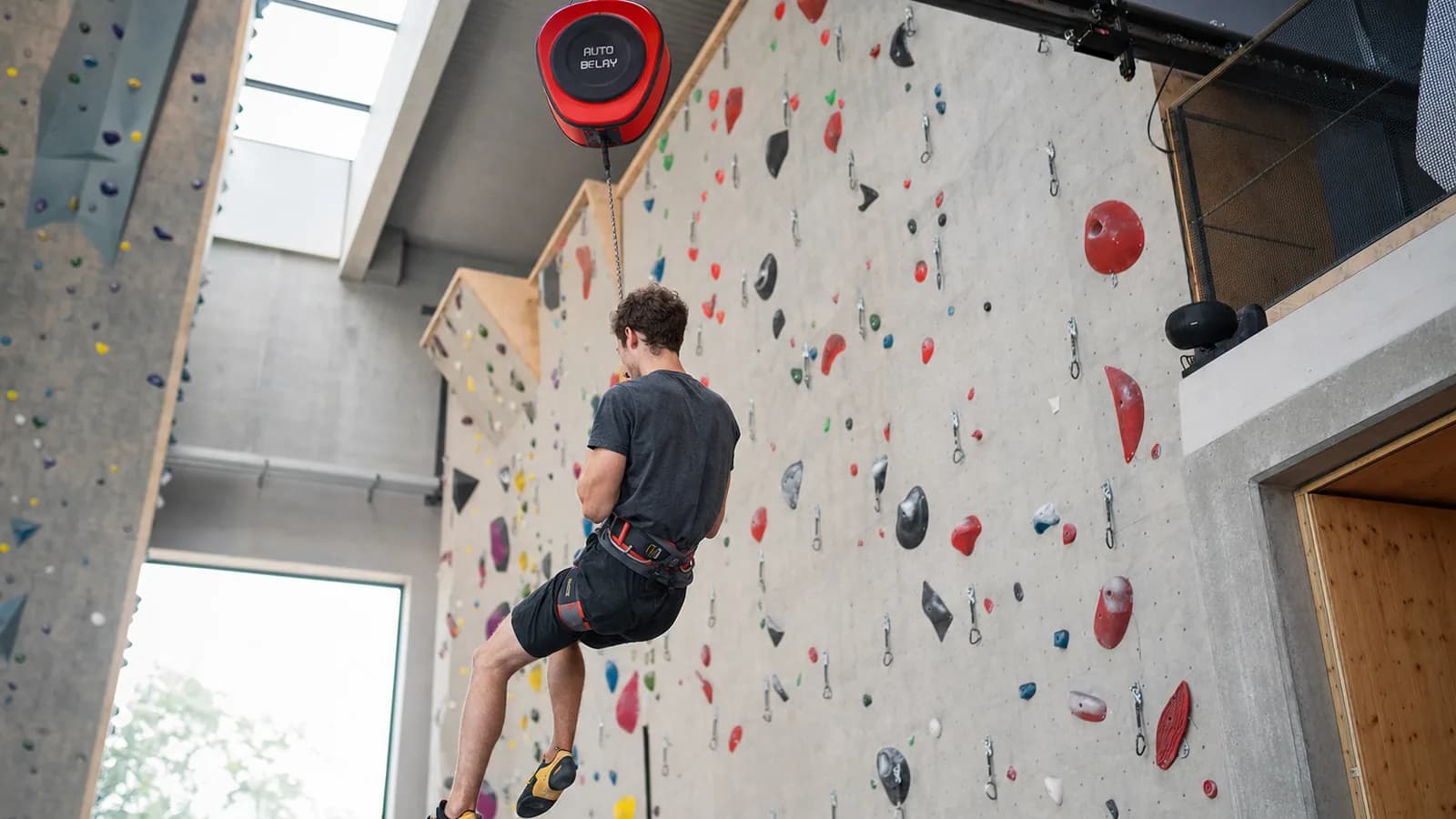 Une grimpeuse en bas d'une voie en salle d'escalade clippe un mousqueton bleu d'autobelay sur son baudrier rouge, prises colorées sur un mur blanc et sangle jaune visible jusqu'en haut