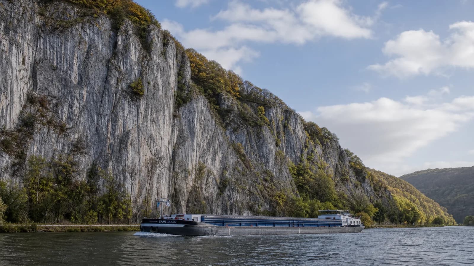 Les Rochers de Freyr vus depuis la rive droite de la Meuse, longue falaise calcaire grise dominant le fleuve, grimpeurs visibles sur les voies du Mérinos, ciel clair de printemps belge