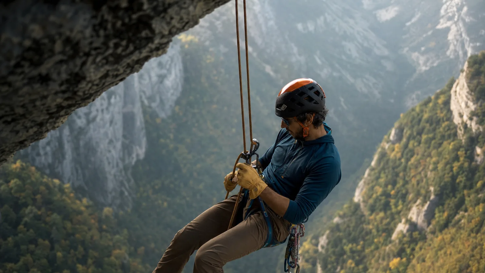 Un grimpeur en plein rappel descend une paroi calcaire ensoleillée en utilisant un descendeur reverso, casque blanc et baudrier noir, autobloquant Machard rouge visible sur la cuisse, deux brins de corde verts dans les mains