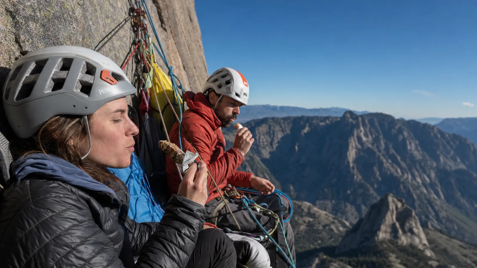 Deux grimpeurs en grande voie installés à un relais suspendu en pleine paroi calcaire ensoleillée, casques rouges et baudriers bleus, matériel ordonné et corde rangée, vide vertigineux en arrière-plan